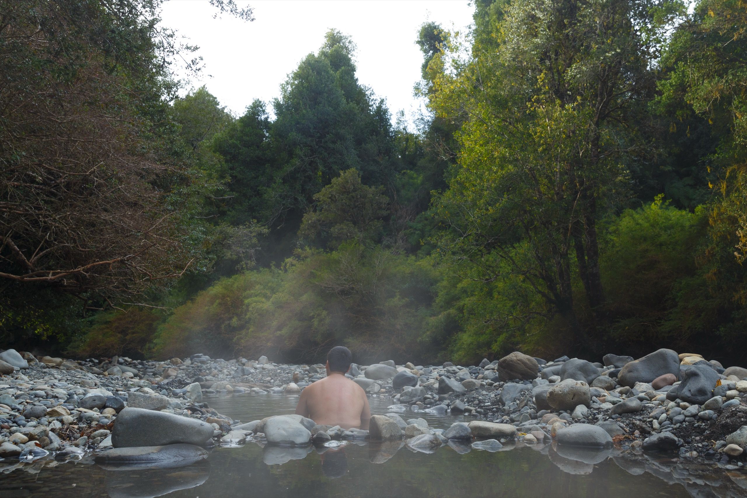 Una persona de espaldas en una poza termal natural entre rocas, con vapor y bosque nativo alrededor, en el Parque Nacional Puyehue